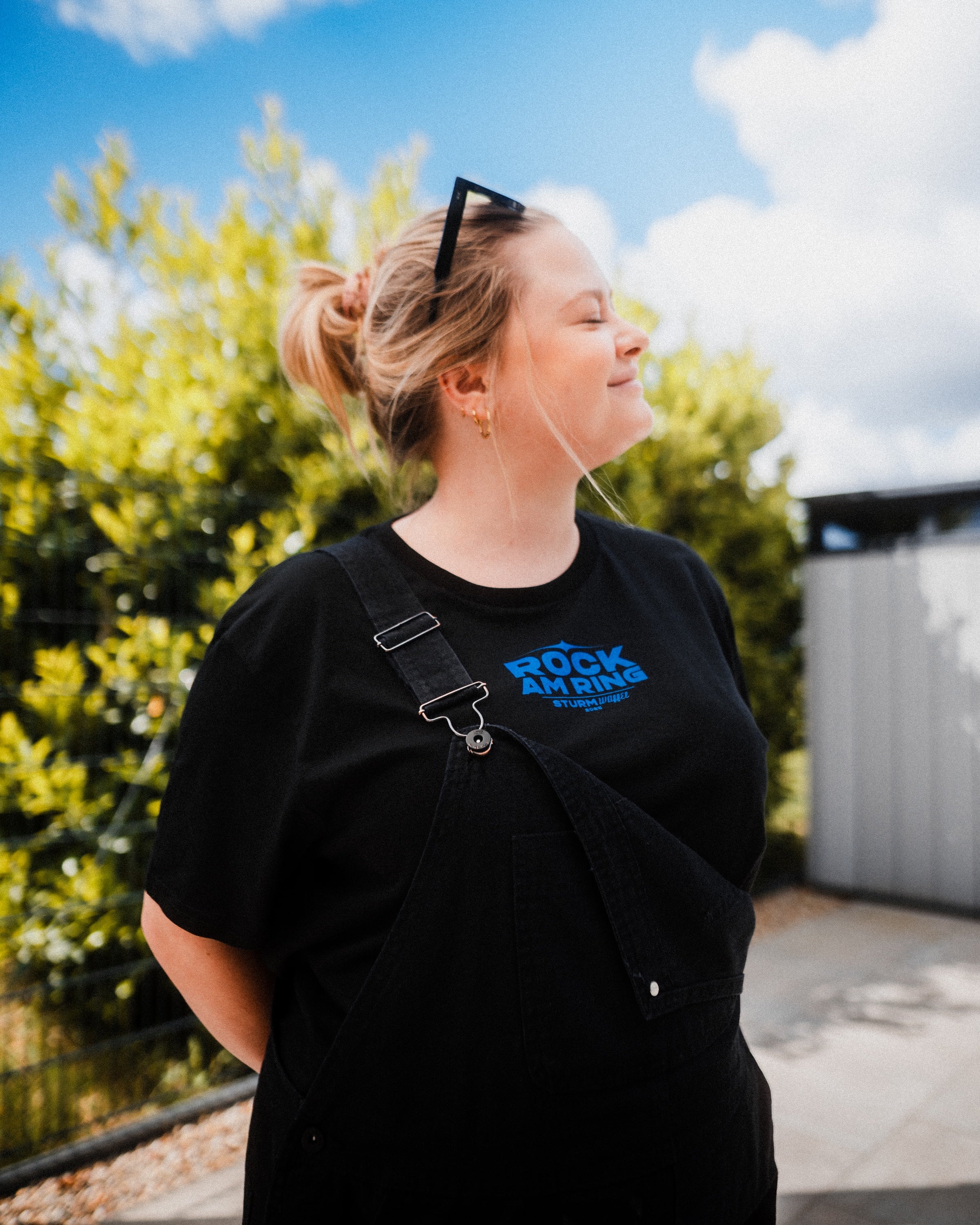 blonde Frau modelt das Sturmwaffel x Rock am Ring - Unisex T-Shirt - Schwarz, kombiniert dazu schwarze Latzhose